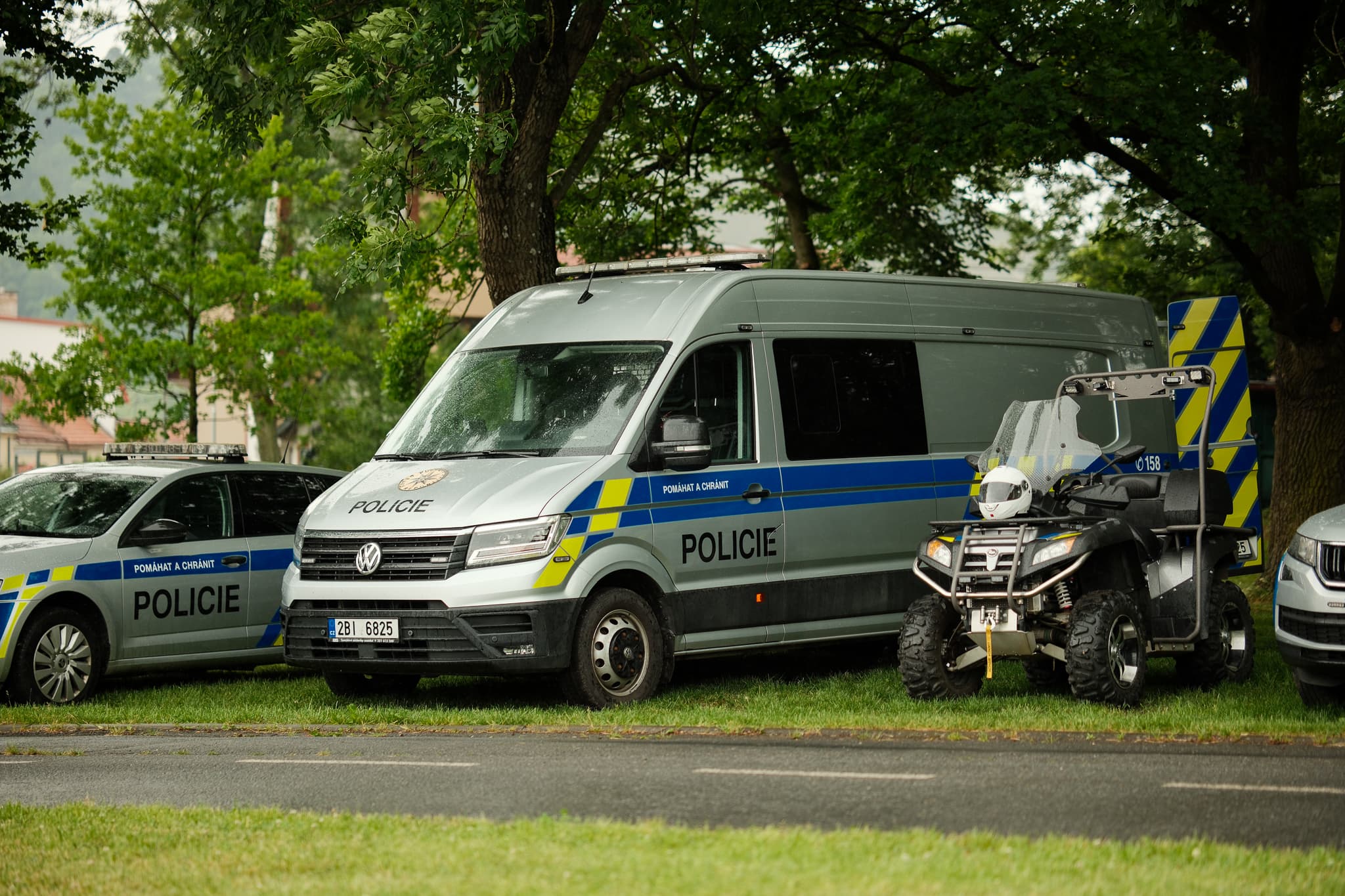 Policejní dodávka, osobní vůz a čtyřkolka zaparkované pod stromy v parku
