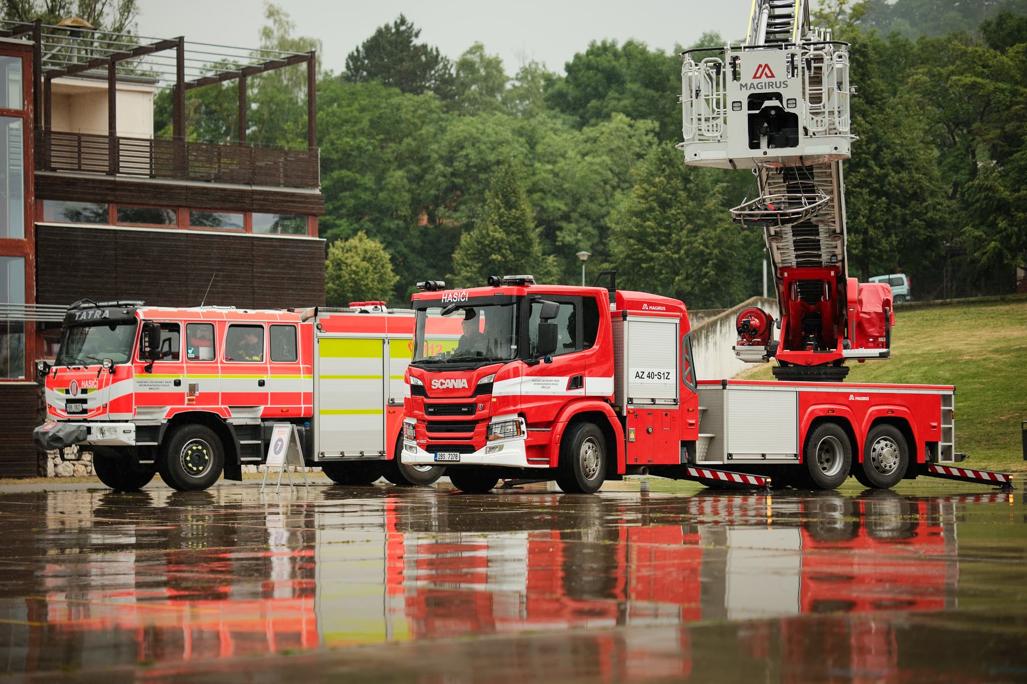 Hasičská technika Tatra a Scania s žebříkem zaparkovaná u budovy za deště