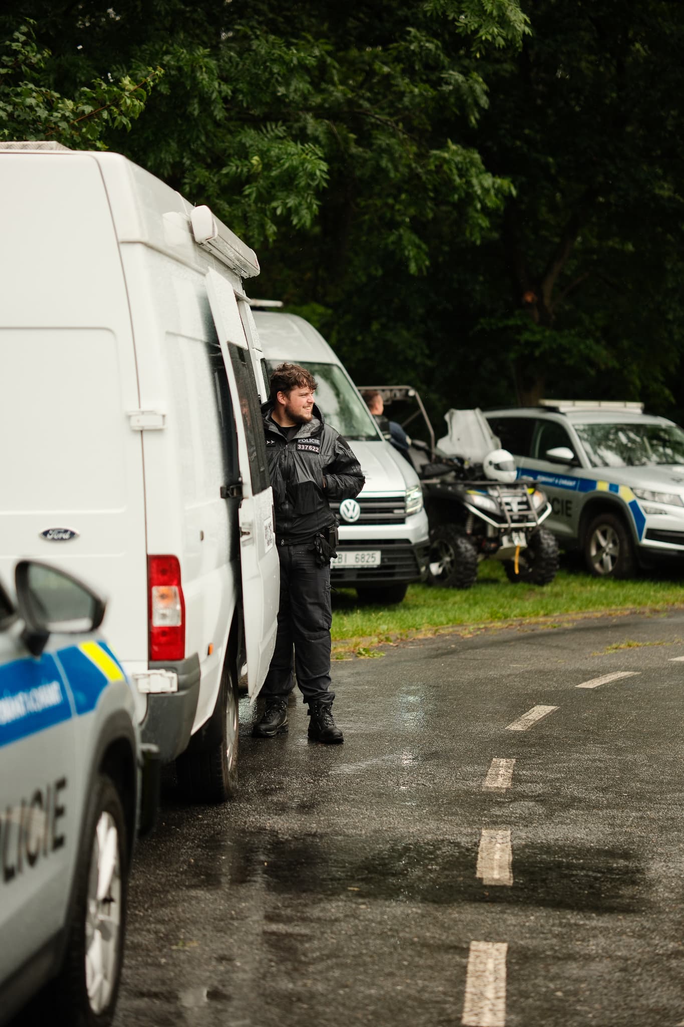 Policista stojí u služebních vozidel zaparkovaných na mokré silnici v parku