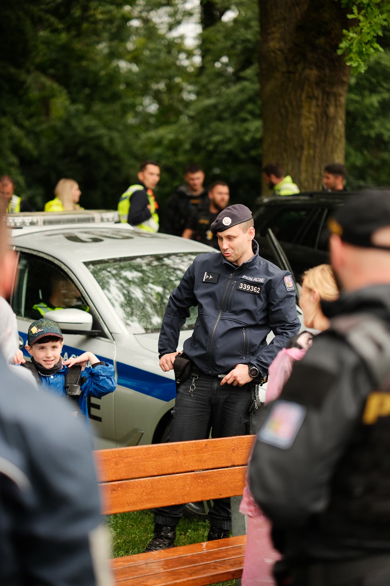 Usměvavý chlapec u policejního auta si zkouší výstroj, kolem stojí policisté