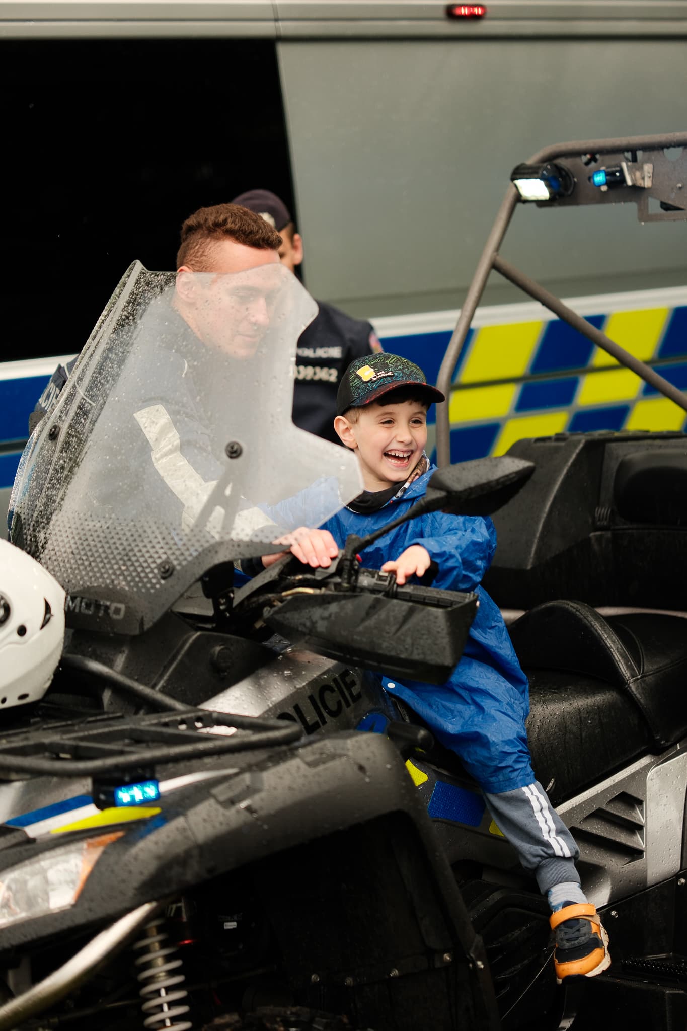 Smějící se chlapec v modré pláštěnce sedí za řídítky policejní čtyřkolky