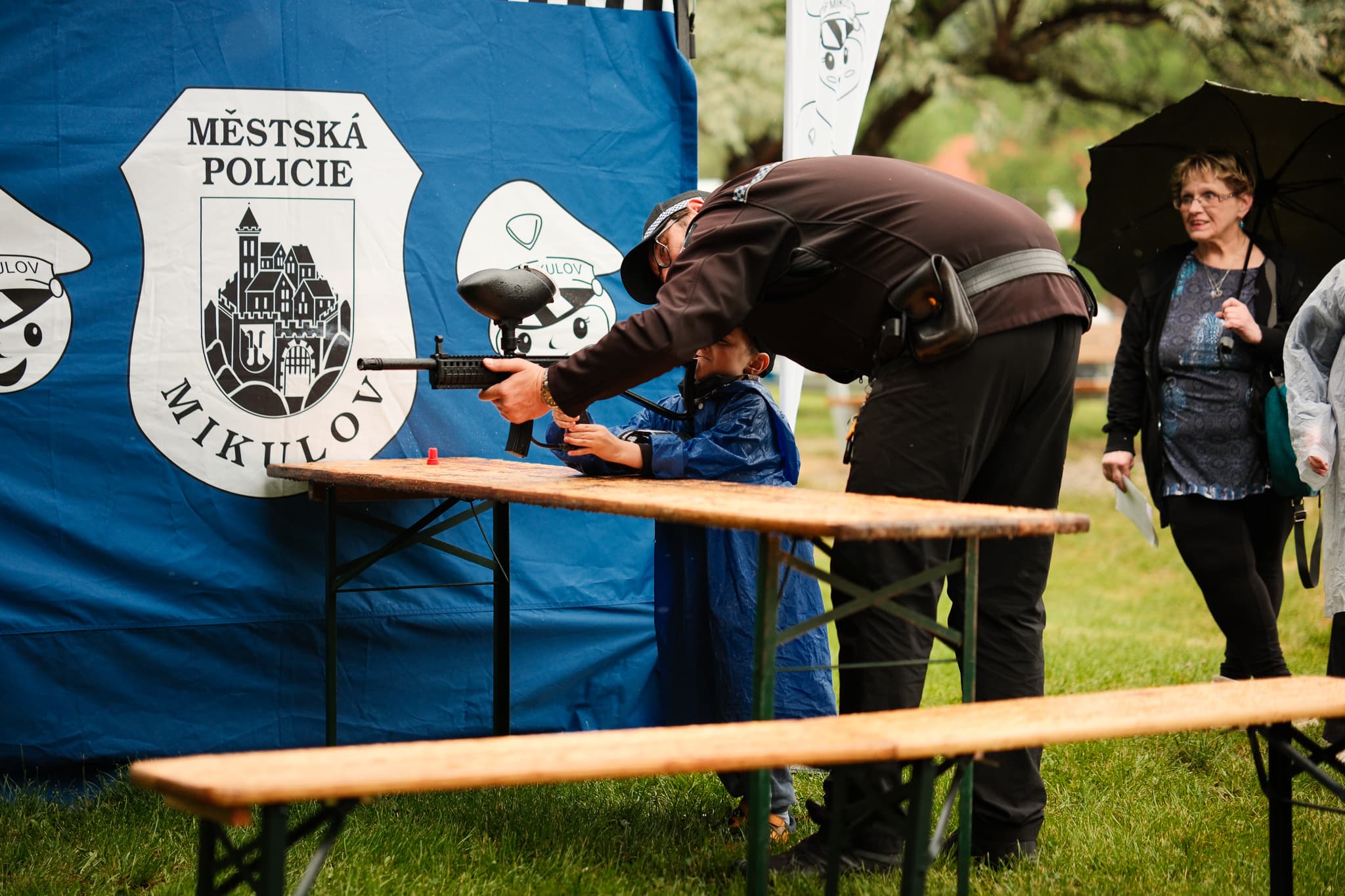 Strážník mikulovské policie pomáhá dítěti mířit paintballovou zbraní u stánku
