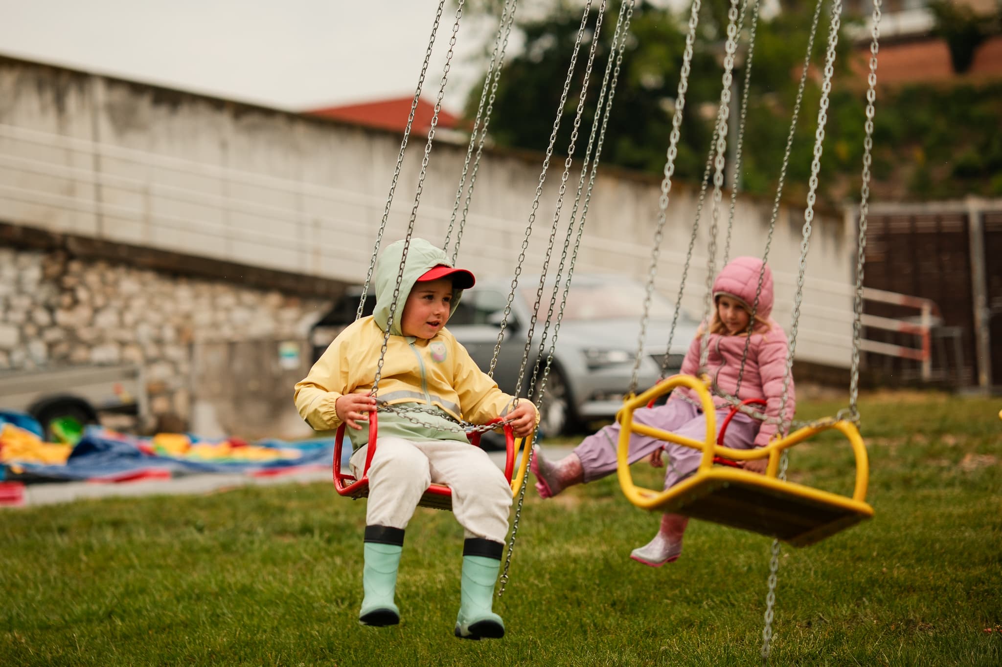 Dvě malé děti v pláštěnkách sedí na houpačkách kolotoče na zatažené louce