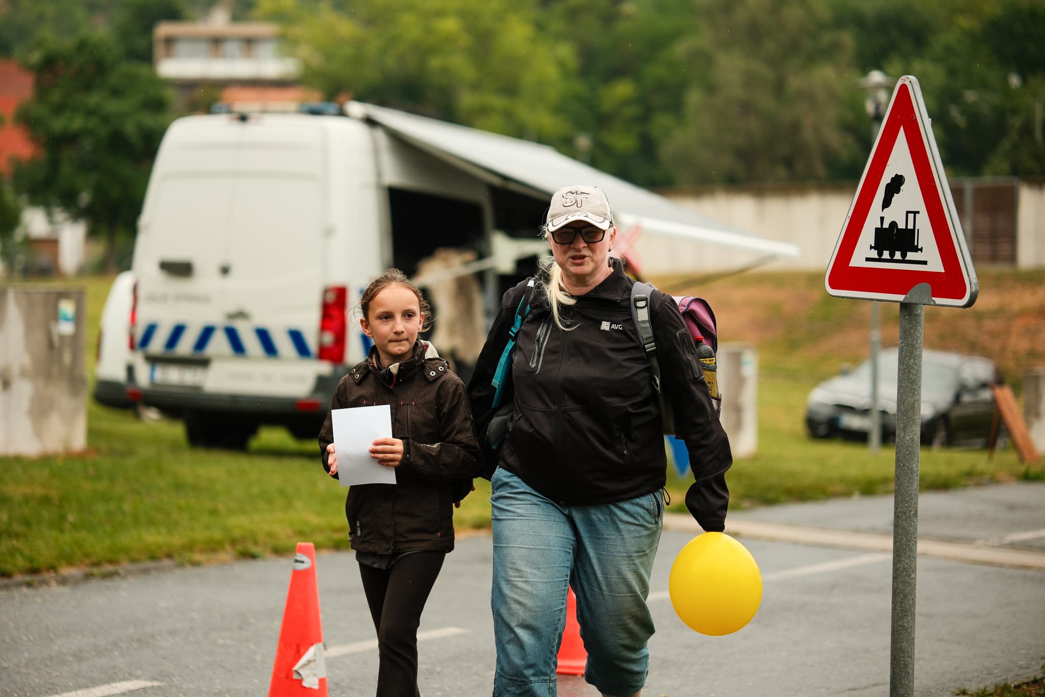 Maminka s dcerou a žlutým balonkem míří přes silnici ke stanovišti