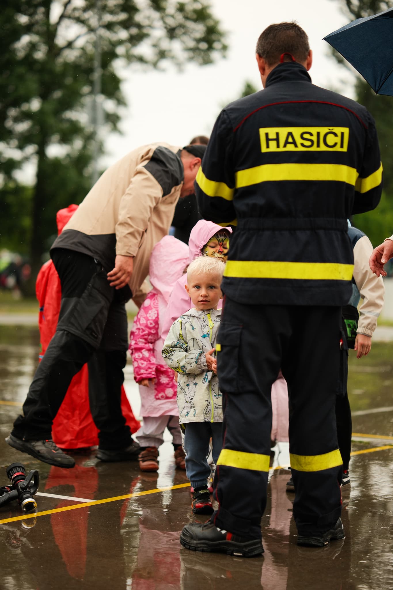 Malý chlapec stojí u hasiče v zásahovém obleku na mokrém asfaltu během Dne dětí