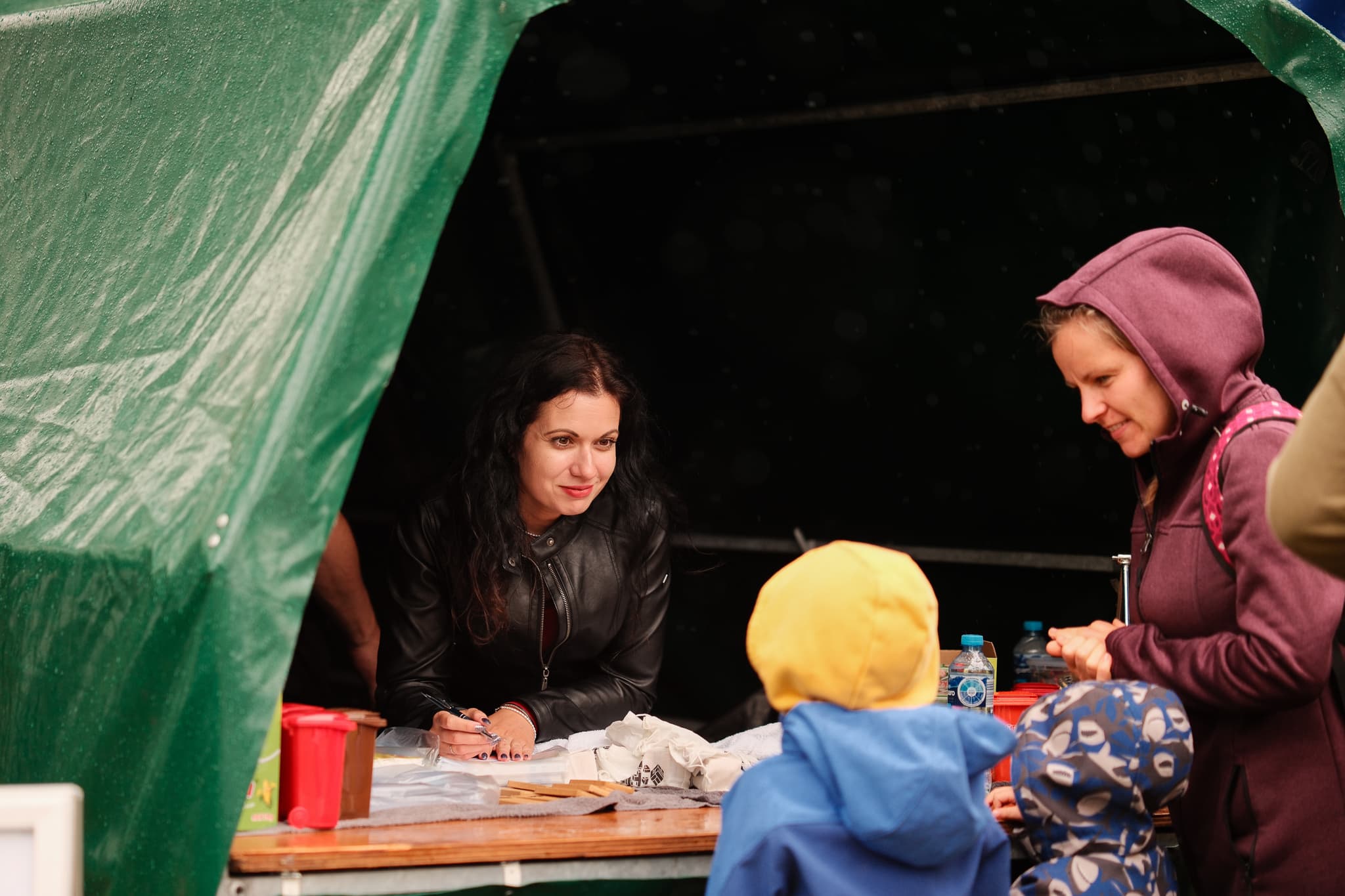 Žena u zeleného stanu vydává dětem materiály na soutěžním stanovišti za deště